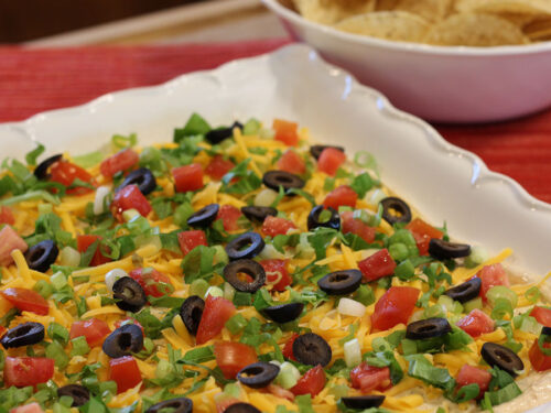 Classic Easy Taco Dip on a serving plate topped with shredded cheddar cheese, lettuce, roma tomatoes, sliced black olives, and green onions.