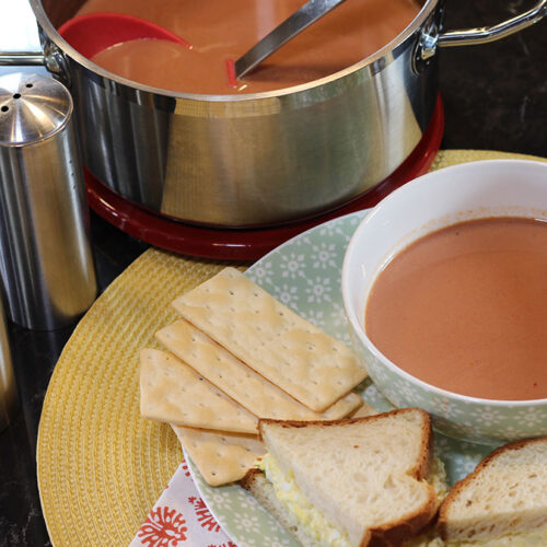 Bowl of homemade tomato juice on a plate with crackers and an egg salad sandwich with a pan of soup in the background