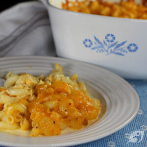 Closeup of baked macaroni and cheese on a plate on a blue mat, white casserole dish with blue flower in background728x728px