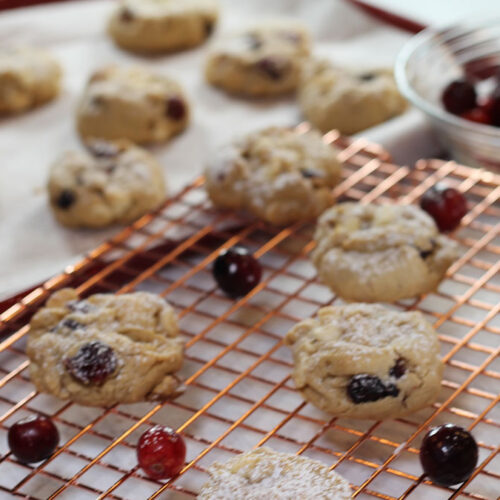 Decadent White Chocolate and Cranberry Oatmeal Cookies with a powdered sugar sprinkle on top. Cooling on a copper wire rack. Background shows a bowl of fresh cranberries and a cookie sheet with baked cookies.