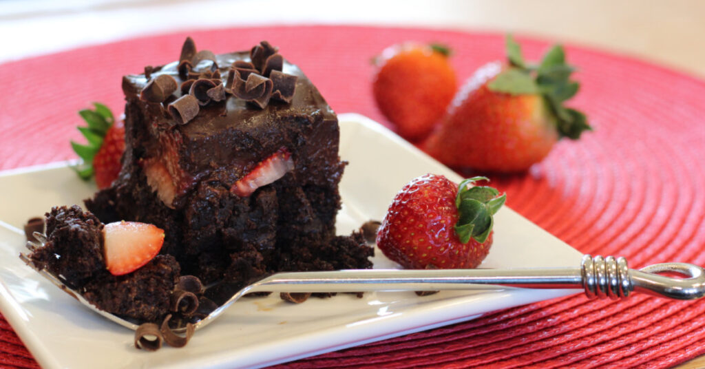 Stylized photo of chocolate ganache and strawberry brownies featuring a brownie on a plate, fork filled with brownie bite and strawberry, fresh strawberries surround the plate.