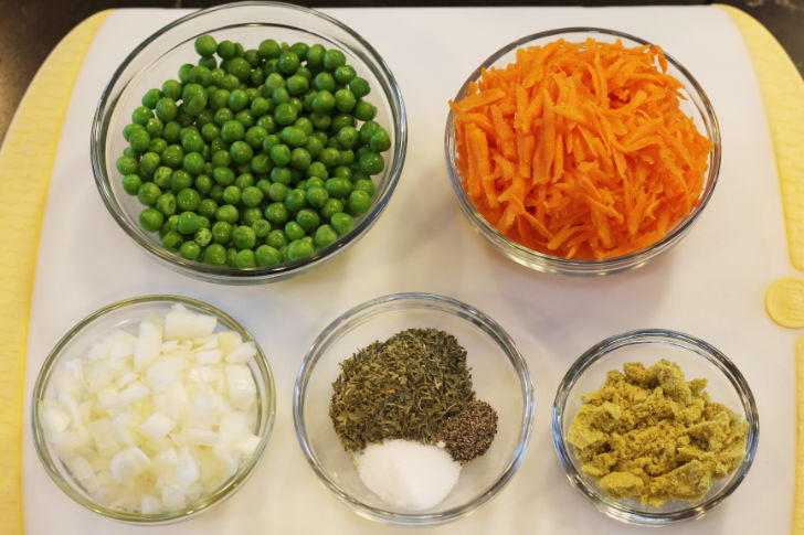 Five small bowls placed on a white board showing the base veggies and aromatics used in the recipe - three small bows contain peas, grated carrot, chopped onions, another containing the parsley flakes, table salt and pepper, and the last bowl with the chicken boullion base.