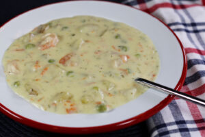 1456x970 px closeup of a bowl of seafood chowder with a spoon in it and a red, white, and blue plaid napkin tucked under the bowl on the right side.