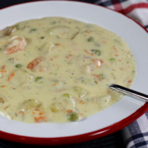 1456x970 px closeup of a bowl of seafood chowder with a spoon in it and a red, white, and blue plaid napkin tucked under the bowl on the right side.