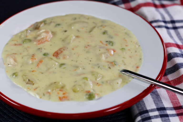 1456x970 px closeup of a bowl of seafood chowder with a spoon in it and a red, white, and blue plaid napkin tucked under the bowl on the right side.