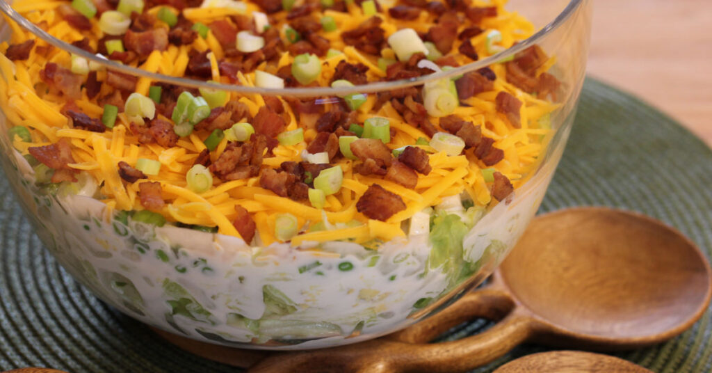 Styled photo showing a clear tall bowl arranged on a green placemat with salad tongs next to it, featuring the seven-layer salad inside.