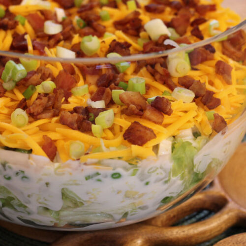 Styled photo showing a clear tall bowl arranged on a green placemat with salad tongs next to it, featuring the seven-layer salad inside.