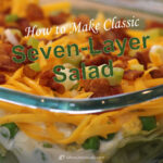 1080x1080 social share image of a styled photo showing a clear dish arranged on a green placemat with seven-layer salad in it.