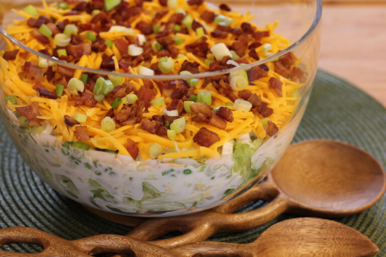 Styled photo showing a clear tall bowl arranged on a green placemat with salad tongs next to it, featuring the seven-layer salad inside.