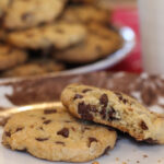 1080x1080 px image showing a closeup of a Chips-Ahoy-style cookie with a bite out of it sitting partially on top of a whole cookie, with a out-of-focus plate of cookies in the background.
