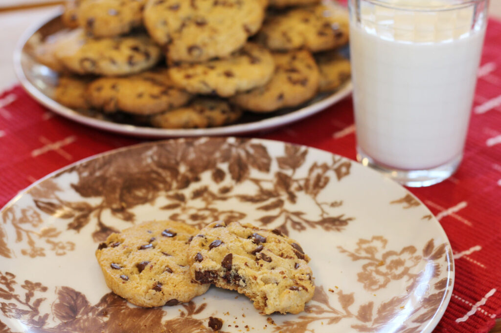 1456x970 px image showing a closeup of a Chips-Ahoy-style cookie with a bite out of it sitting partially on top of a whole cookie, with a out-of-focus plate of cookies in the background and a glass of milk, with everything sitting on a red placemat.