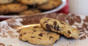1200x628 px image showing a closeup of a Chips-Ahoy-style cookie with a bite out of it sitting partially on top of a whole cookie, with a out-of-focus plate of cookies in the background.