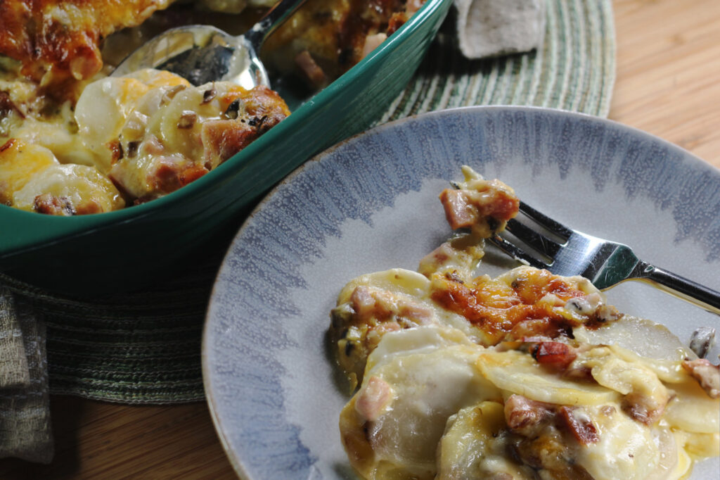 Closeup photo of Cheesy Scalloped Potatoes with Ham and Mushrooms on a blue plate with a small bite of food on fork in the lower right corner. A green casserole dish is pictured in the upper left with a spoon scooping the scalloped potatoes. The styled photo shows both dishes on a blue-green colored placemat.