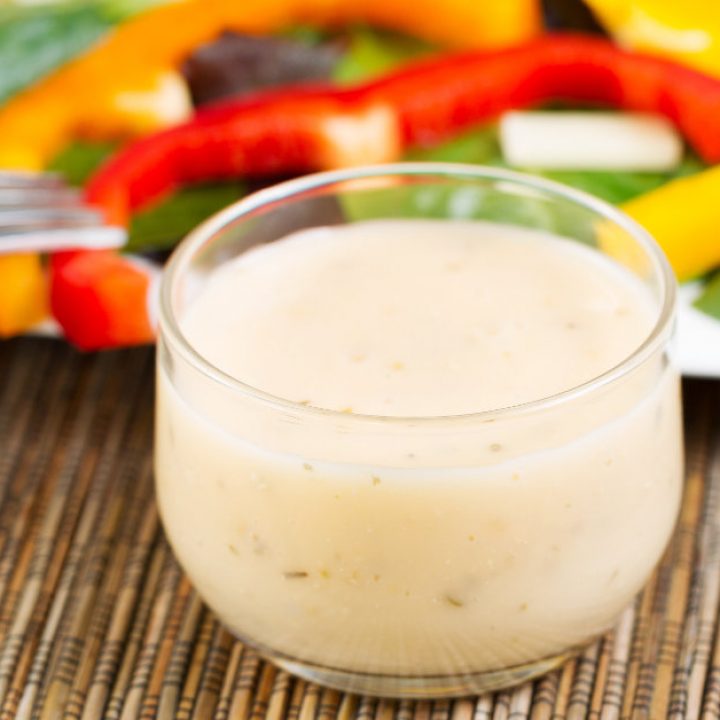 Horizontal photo of a small glass bowl filled with creamy Italian dressing with salad on white plate in background