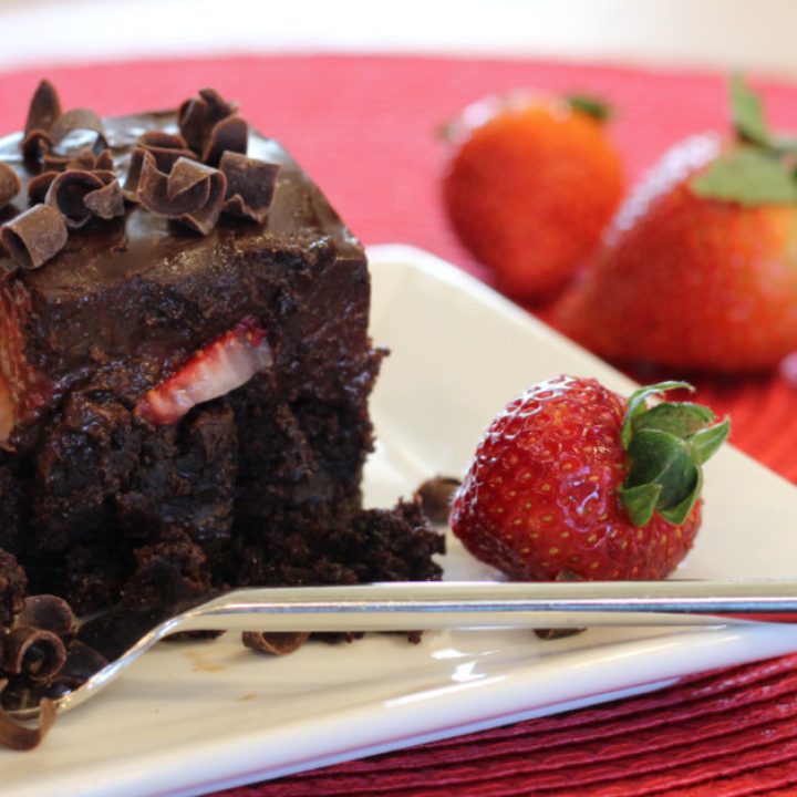 Stylized photo of chocolate ganache and strawberry brownies featuring a brownie on a plate, fork filled with brownie bite and strawberry, fresh strawberries surround the plate.