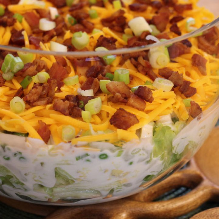 Styled photo showing a clear tall bowl arranged on a green placemat with salad tongs next to it, featuring the seven-layer salad inside.
